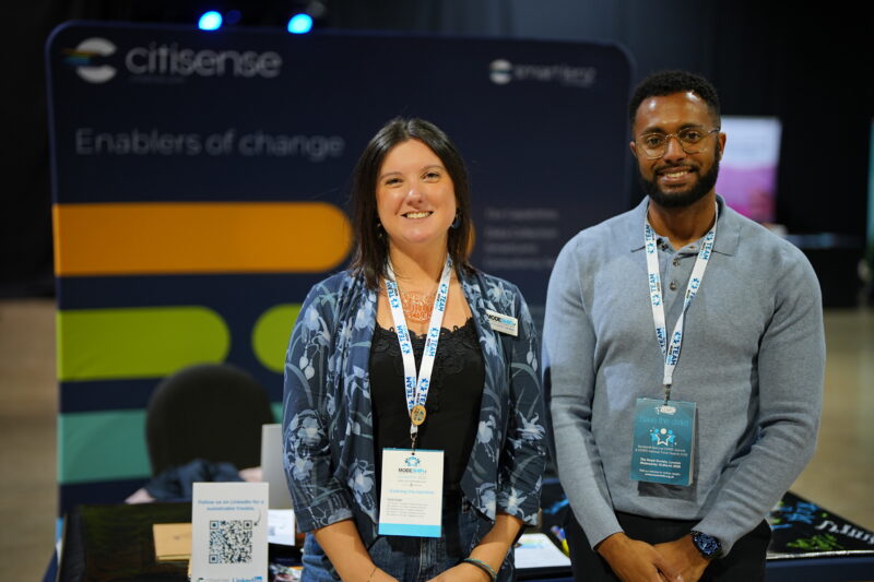 Two individuals stand in front of a "Citizense Enablers of Change" backdrop at what appears to be a conference or trade show booth.