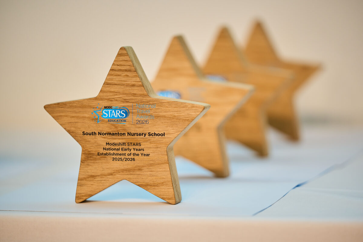 A row of four wooden star-shaped awards with engraved text sits on a light blue surface. The closest star is in sharp focus and prominently displays "South Normanton Nursery School" along with "Modeshift STARS National Early Years Establishment of the Year 2025/2026" and a logo. The other three stars are progressively blurred in the background.