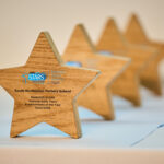 A row of four wooden star-shaped awards with engraved text sits on a light blue surface. The closest star is in sharp focus and prominently displays "South Normanton Nursery School" along with "Modeshift STARS National Early Years Establishment of the Year 2025/2026" and a logo. The other three stars are progressively blurred in the background.
