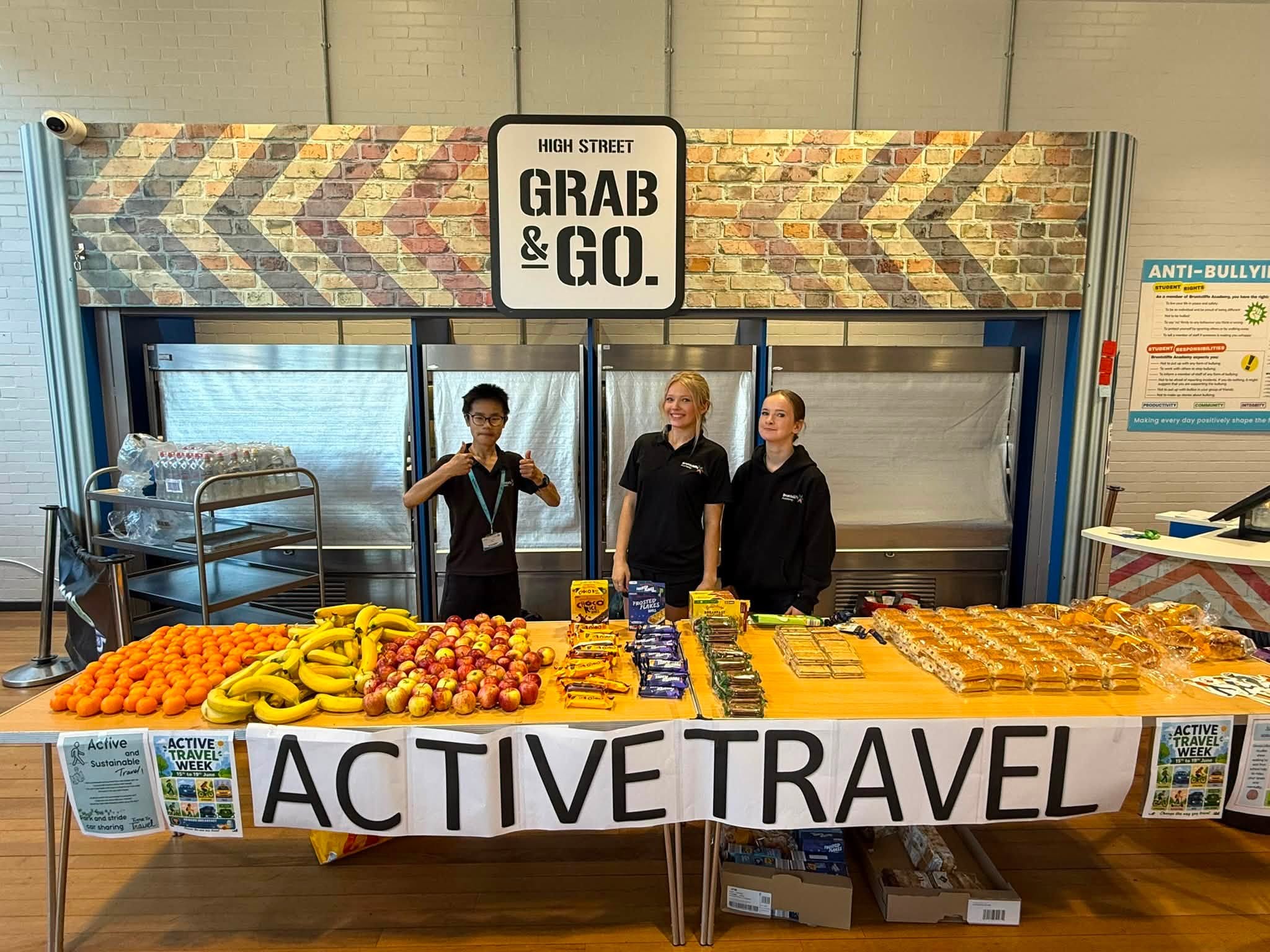A stall promoting "Active Travel Week" displays fresh fruit such as oranges, bananas, and apples alongside packaged snacks and baked goods. Three individuals stand behind the stall, with two young women in black hoodies and a young person in a dark t-shirt wearing a lanyard. A sign above them reads "HIGH STREET GRAB & GO."