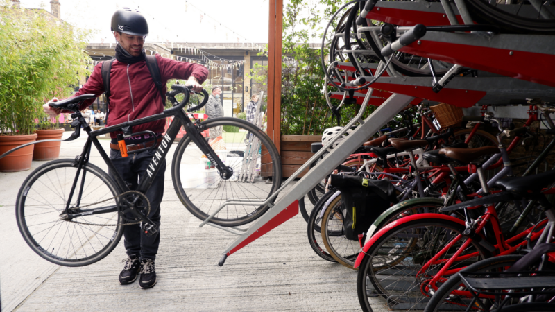 Man in maroon jacket and helmet holding a black bicycle with a chain lock on the frame, standing next to a multi-level bike rack filled with many bicycles. Turvec photo credit.