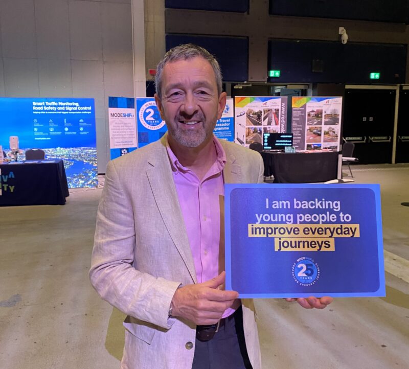 A man in a light linen jacket and pink shirt holds a blue sign that reads "I am backing young people to improve everyday journeys" with a 25th anniversary logo.