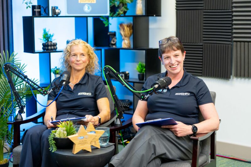Two women in matching dark blue polo shirts with a white logo sit in armchairs in a recording studio, each with a microphone and a clipboard.