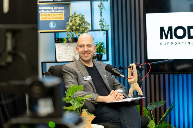A bald man in a gray suit jacket smiles while sitting in a chair with a microphone in front of him and a logo on a screen behind him.