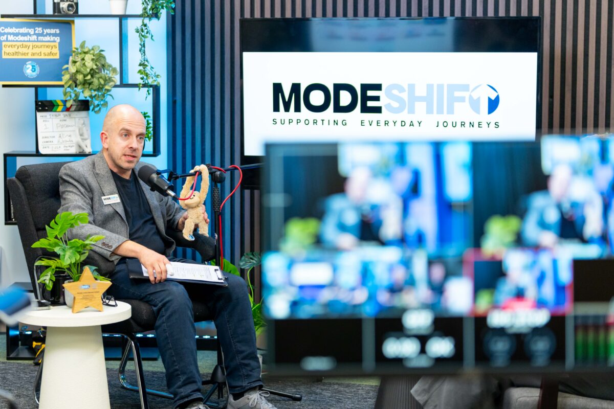 A man wearing a grey blazer sits in a chair holding a stuffed toy and a clipboard, speaking into a microphone in front of a screen displaying the "MODESHIFT SUPPORTING EVERYDAY JOURNEYS" logo.
