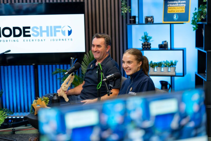 Two people sit at a table with microphones and a stuffed monkey, smiling and looking to the side, in front of a large screen displaying the "ModeShift" logo.