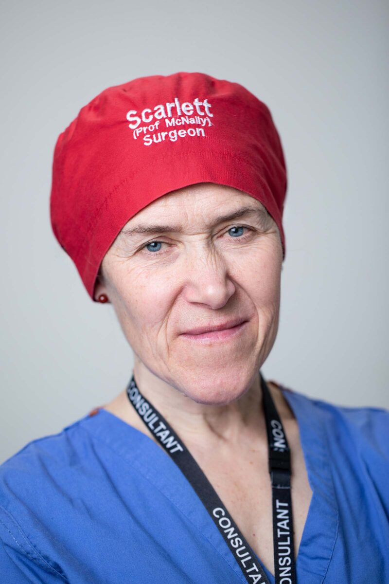 A professional portrait of a surgeon wearing a red surgical cap embroidered with "Scarlett (Prof McNally) Surgeon" and a blue scrubs top with a lanyard reading "CONSULTANT."