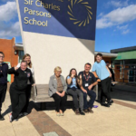 A group of people, including students and adults, are gathered outdoors in front of a sign that reads "Sir Charles Parsons School". Several individuals are smiling and giving thumbs up.