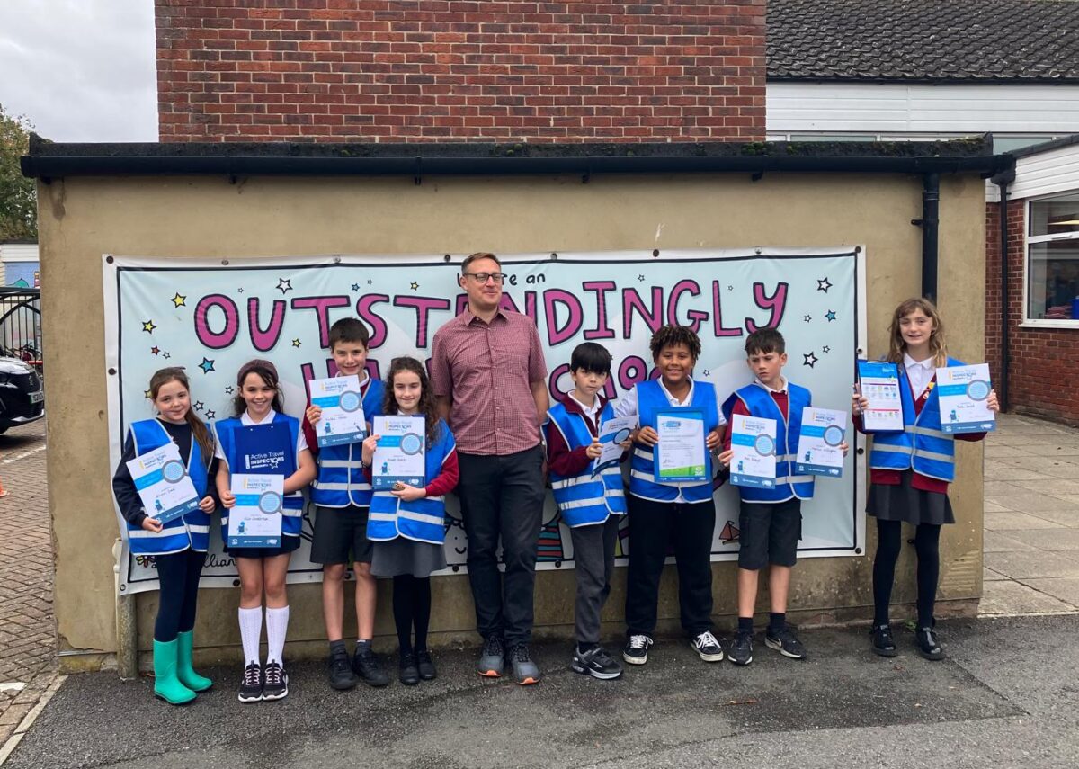 Children in bright blue safety vests and a man stand in front of a banner that reads "Outstandingly" with certificates in their hands.