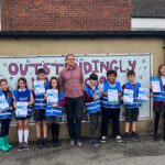 Children in bright blue safety vests and a man stand in front of a banner that reads "Outstandingly" with certificates in their hands.
