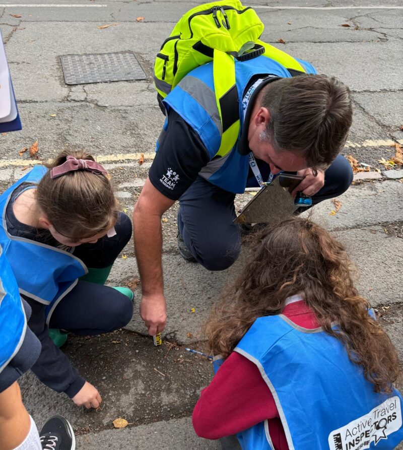 An adult wearing a bright yellow backpack and blue reflective vest crouches down to examine the ground with two children wearing similar blue vests, who are also looking at the pavement.