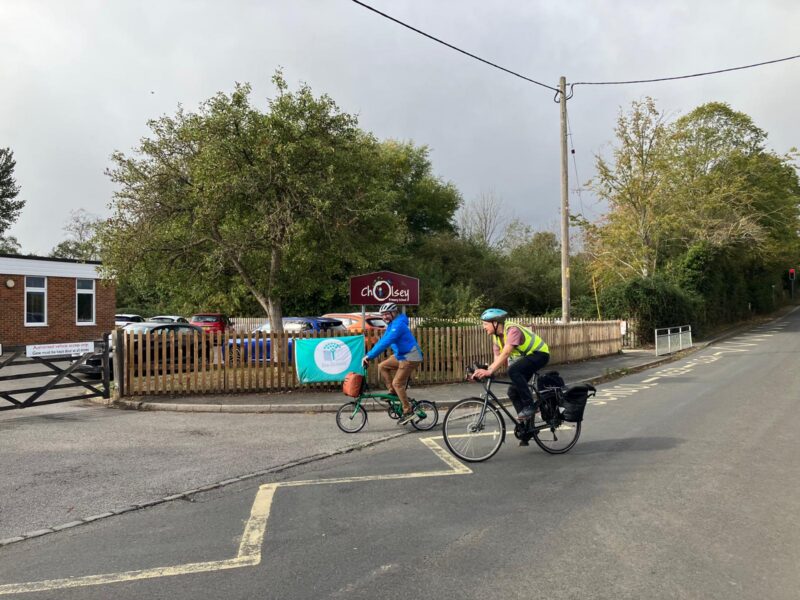 Two cyclists ride past a school entrance with a wooden fence and a banner.