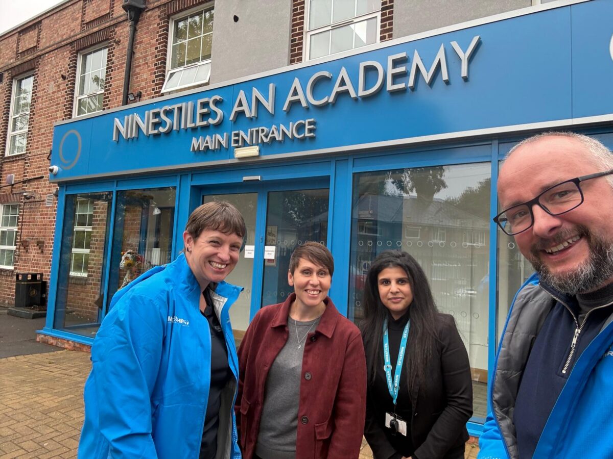 Three individuals stand outside the main entrance of Ninestiles An Academy, a building with a blue facade and large windows.