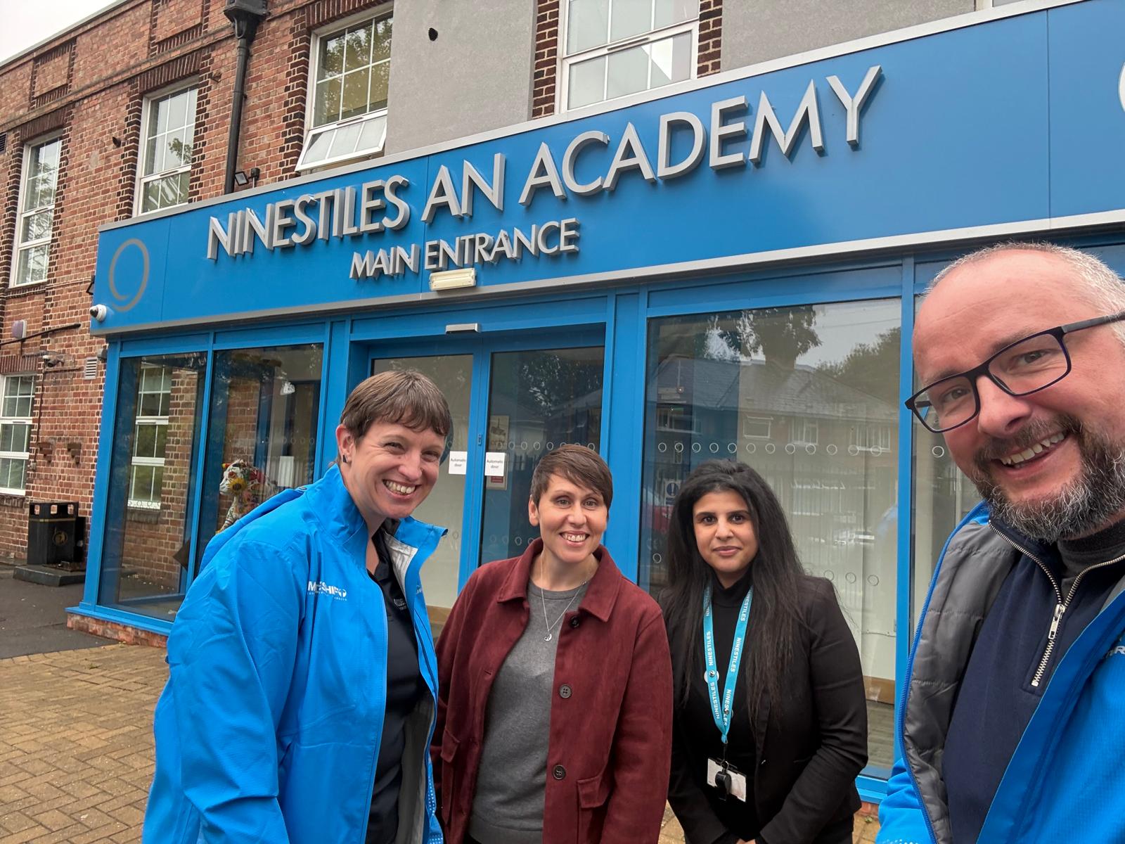 Three individuals stand outside the main entrance of Ninestiles An Academy, a building with a blue facade and large windows.