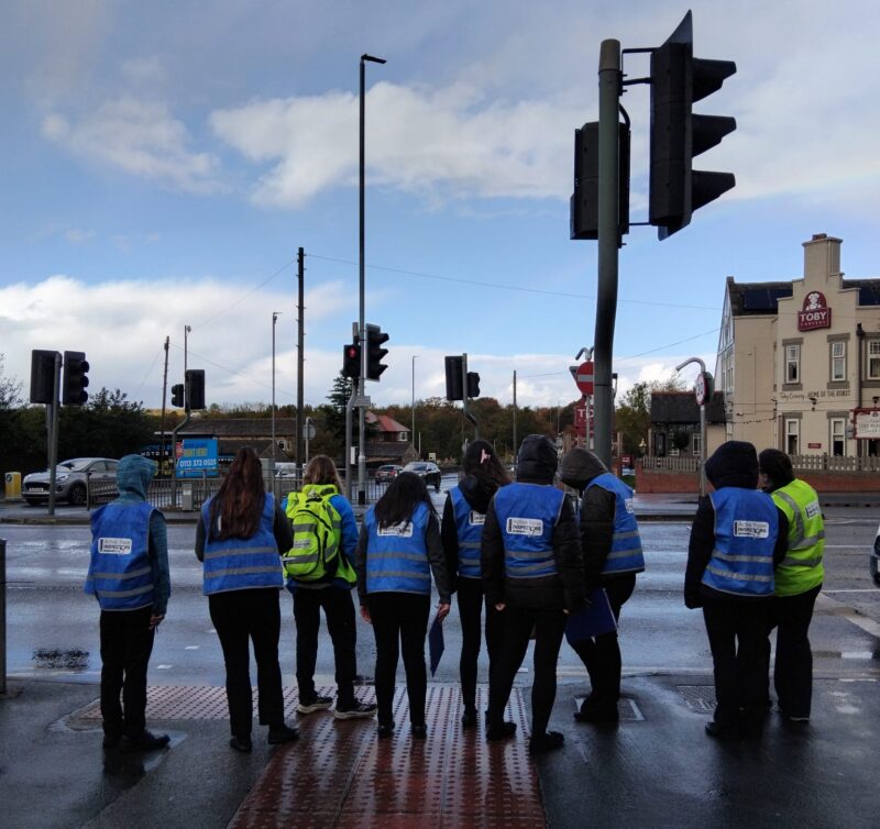 A group of young people wearing bright blue safety vests stand at a wet pedestrian crossing, waiting for the traffic lights to change.