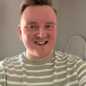 A man smiling indoors, wearing a light green and white striped shirt, standing in front of a cream wall with a mirror in the background.
